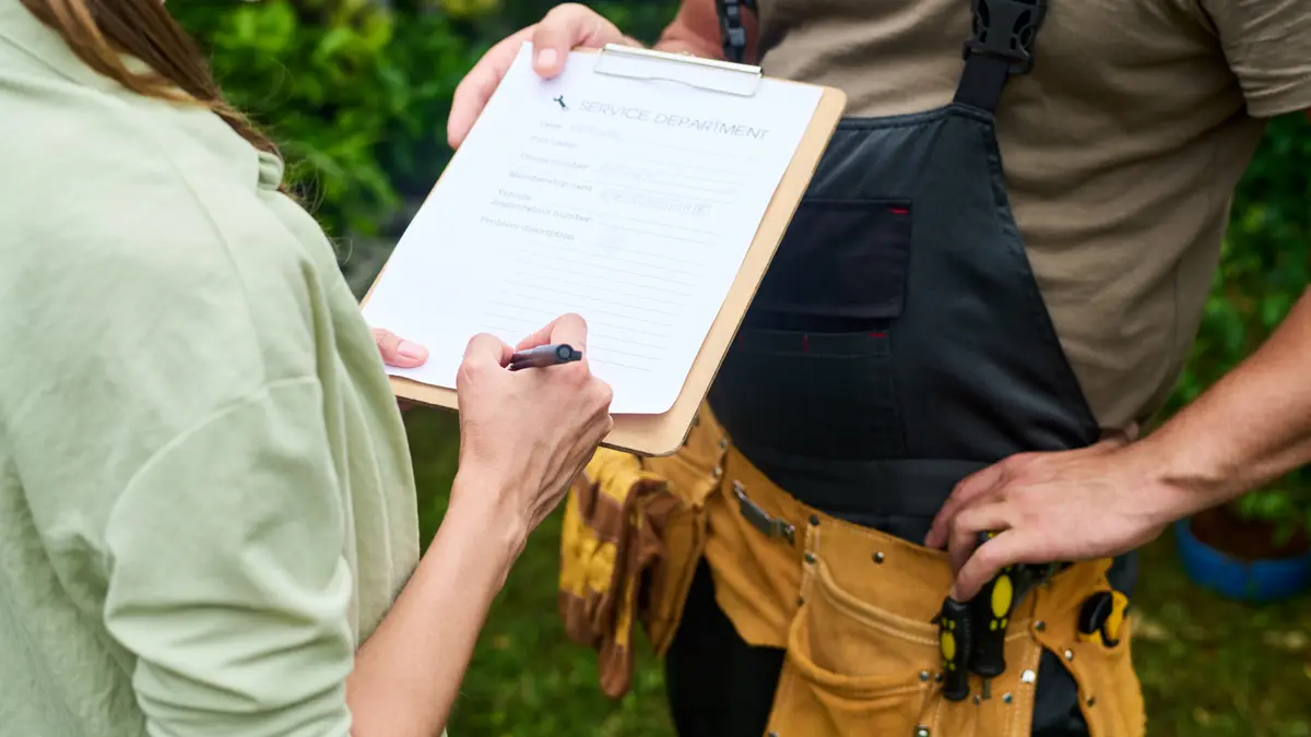 Clienta firma el contrato de obra en la tablilla que le sostiene un contratista con cinturón de herramientas