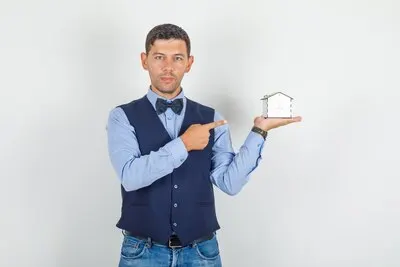 Young man in suit, jeans pointing finger at house model and looking hopeful