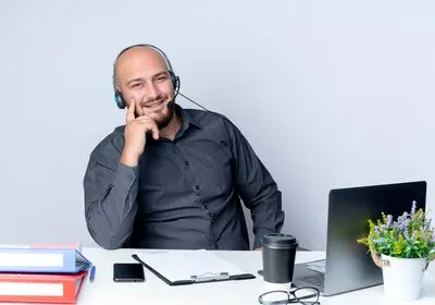 Smiling young bald call center man wearing headset sitting at desk with work tools putting finger on cheek isolated on white wall