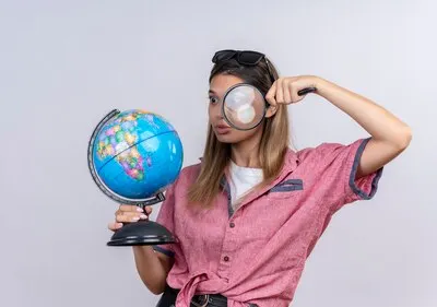 A shocked young woman wearing red shirt in sunglasses holding a globe while looking at it with magnifying glass on a white wall