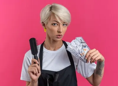 Confident young beautiful female barber in uniform holding comb with cash isolated on pink wall