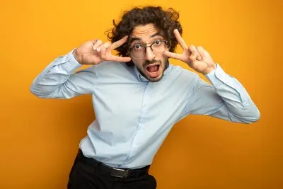 Impressed young handsome caucasian man wearing glasses looking at camera keeping hands near head doing peace signs isolated on orange background