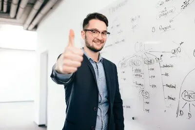 Young dark-haired man in glasses is standing near whiteboard in office. He wears blue shirt and dark jacket. He is smiling and showing a sign  to the camera.