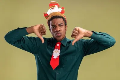 African american man with funny santa rim and red tie  showing thumbs down with angry face standing over green wall