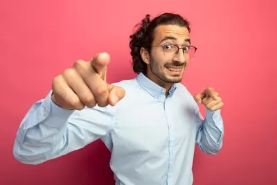 Impressed young handsome caucasian man wearing glasses looking at camera smiling and doing you gesture isolated on crimson background