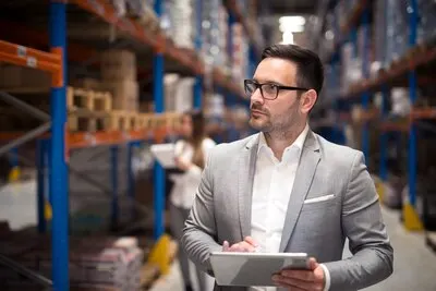 Portrait of successful businessman manager CEO holding tablet and walking through warehouse storage area looking towards shelves