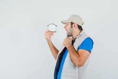 Young man looking at house model in t-shirt, jacket, cap and looking amazed , front view.