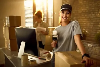 Happy courier using tape dispenser while packing boxes for a delivery and looking at the camera