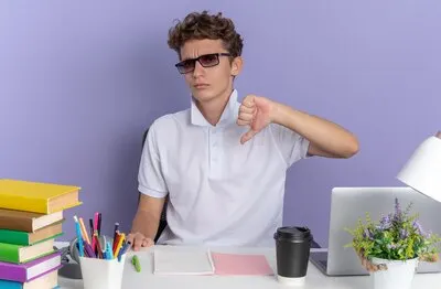 Student guy in white polo shirt wearing glasses sitting at the table with books looking at camera with serious face pointing at himself over blue background