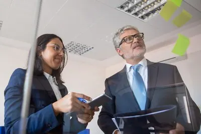 Focused colleagues reading stickers on glass wall and standing. Content young Latin businesswoman holding tablet, bearded man holding clipboard. Management, business and partnership concept