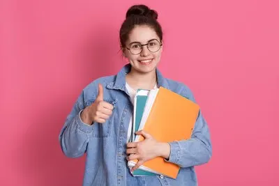 young student woman wearing denim jacket and eyeglasses, holding colorful folders and showing thumb up on pink