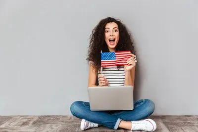 Gorgeous female with beautiful smile sitting in lotus pose with silver computer on legs demonstrating american flag over grey wall