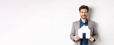 Real estate and insurance concept salesman in grey suit showing paper house cutout selling property