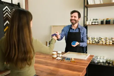Young female client is handing the smiling salesman her credit card to pay for her art supplies in the store