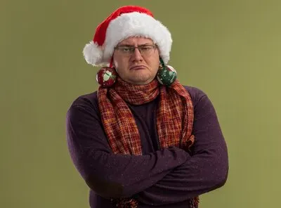 confident adult man wearing glasses and santa hat with scarf around neck standing with closed posture looking at camera with christmas baubles hanging from his ears isolated on olive green background