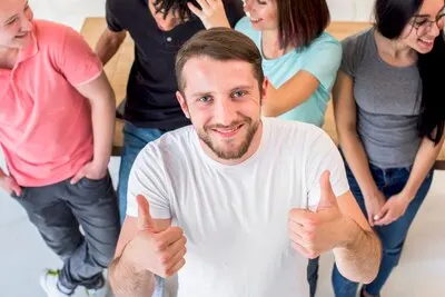 Happy young man standing with friends showing thumbup gesture looking at camera