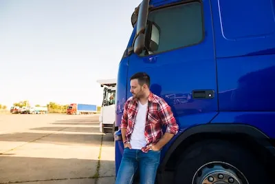 Camionero apoyado en la cabina de un camión azul en un aparcamiento de camiones
