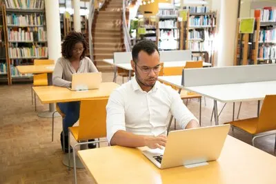 Adultos hacen un examen en computadora portátil en las mesas de una biblioteca